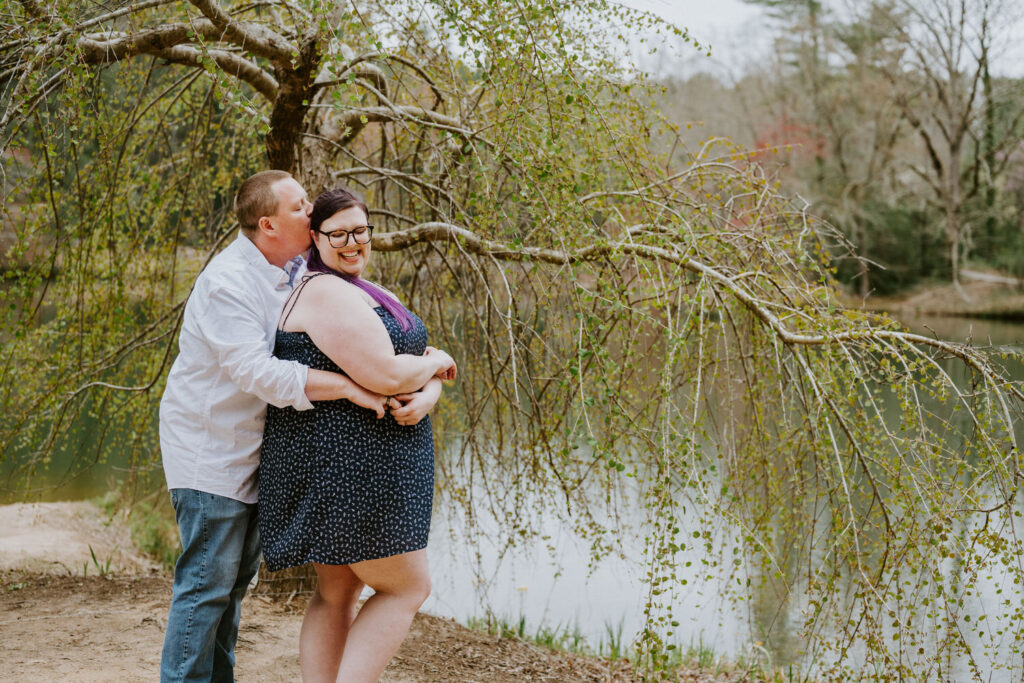 Couple standing by a pond behind each other with their arms wrapped around each other. 
