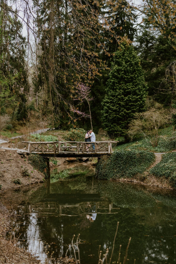 Couple standing on a bridge over a pond with their image reflecting in the water below.