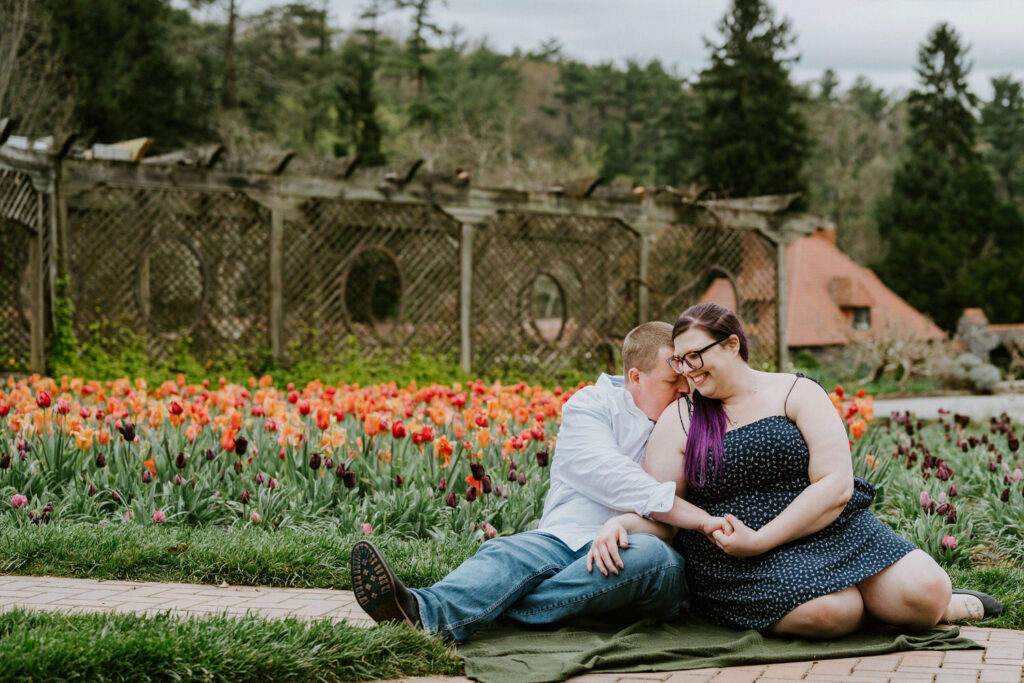 Two people sitting down in a colorful garden while one kisses the other's shoulder.