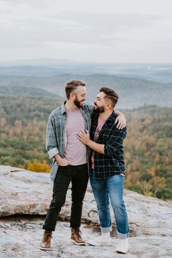 A couple standing on top of a rock overlooking a mountain with their arms around each other.