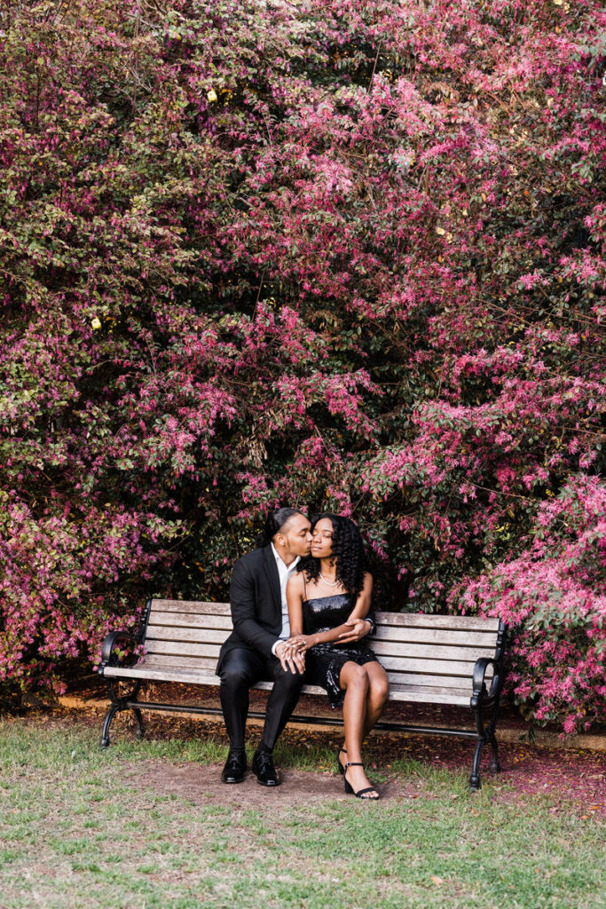 A couple sitting on a wooden bench in front of a large flower bush as he leans in and kisses her cheek. 