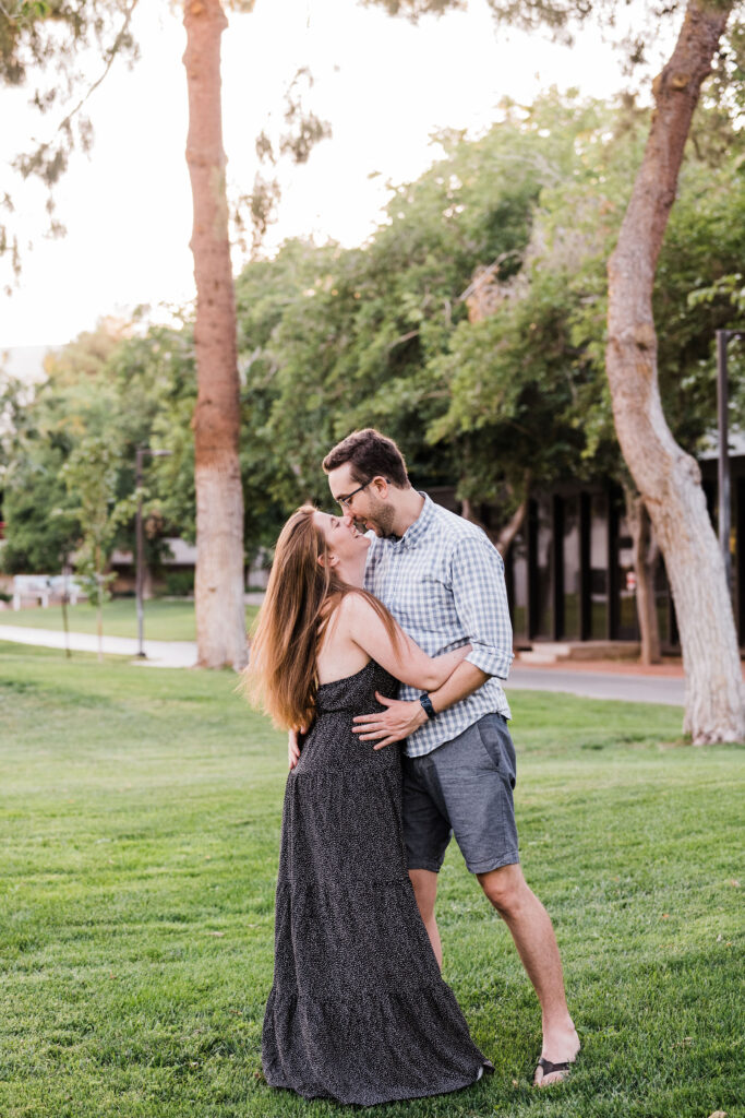 A couple with their arms around each other's waists as they lean in for a kiss while standing in a small grassy area with trees around them. 