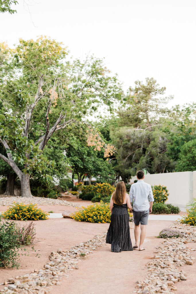 A couple holding hands and walking on a dirt path lined with rocks on either side in a desert garden. 