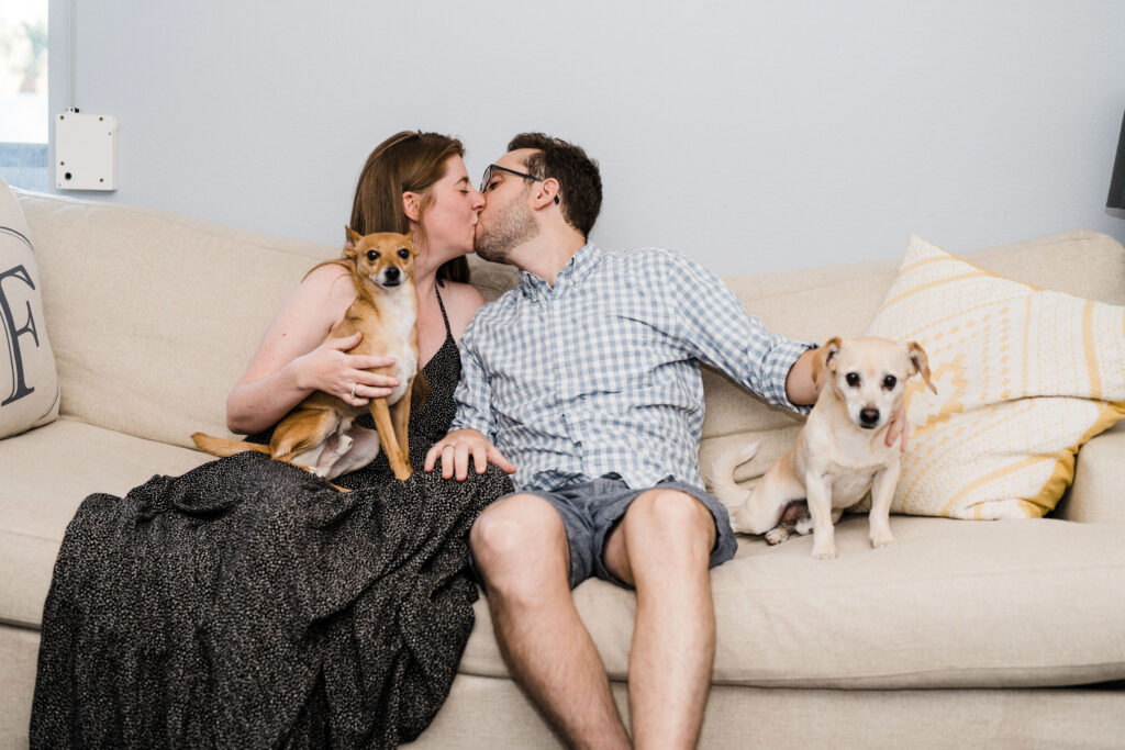 A couple sitting on a couch next to each other kissing while one is holding a dog on their lap and the other has their hand on a dog next to them. 