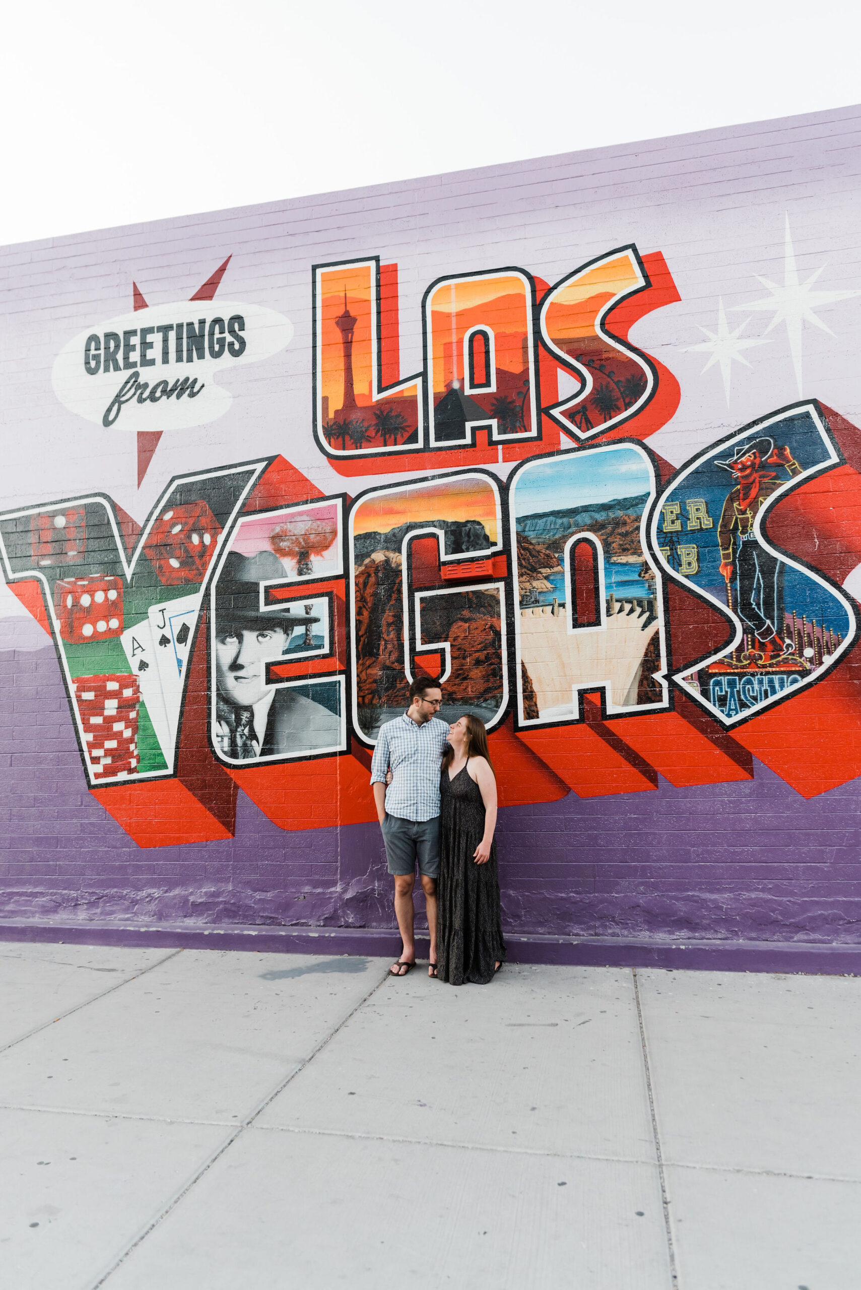 Las Vegas engagement photos of a couple with their arms around each other smiling while standing in front of a large mural that reads "Greeting from Las Vegas".