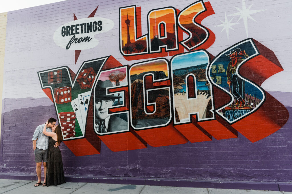 A couple kissing while standing to the left side of a large mural on the side of a building that reads "Greetings from Las Vegas". 