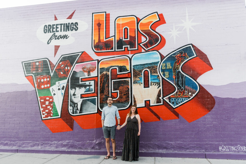 A couple holding hands and standing in front of a wall with a large, colorful mural that says "Greetings from Las Vegas". 