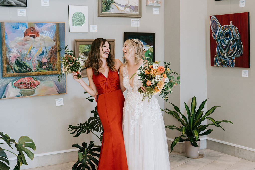 A bride and her bridesmaid standing next to each other with their arms around each other laughing as they hold bouquets of flowers. 