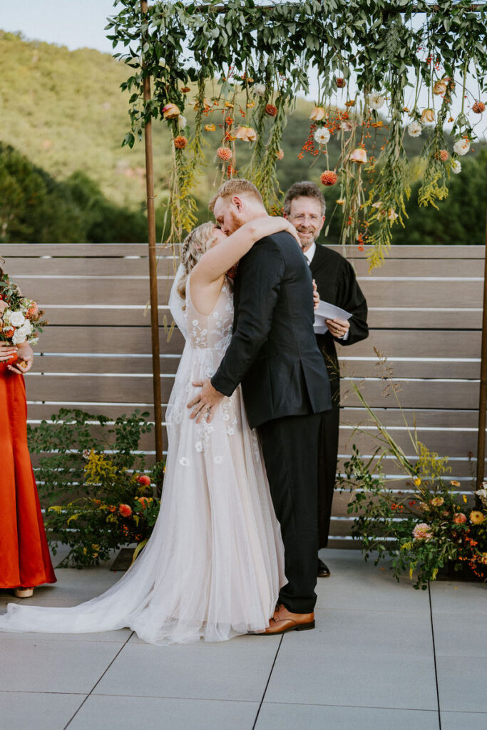 A bride and groom during their first kiss with their officiant behind them. 