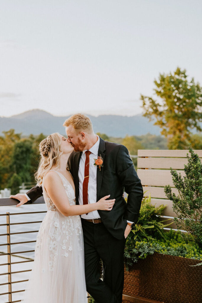 A bride and groom kissing while standing on a rooftop patio with a view of the mountains. 