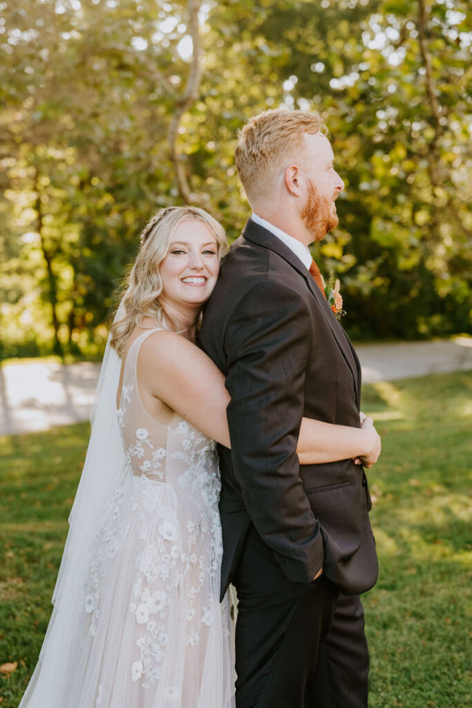 A bride smiling while she wraps her arms around her groom from behind. 