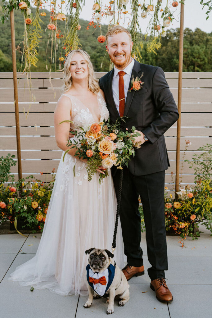 A bride and groom standing together smiling after getting married with their pug sitting at their feet. 