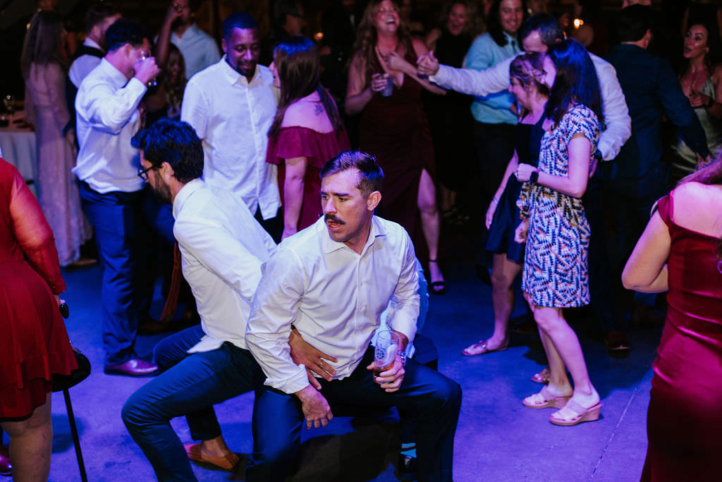 Two wedding guests dancing back to back as they squat down during a reception. 