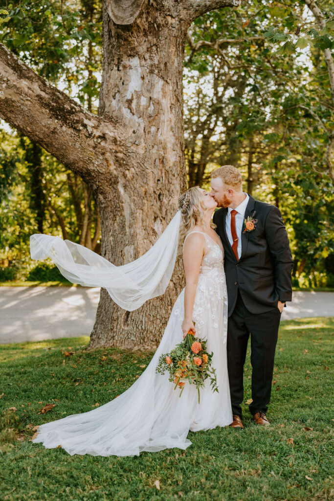 A bride and groom kissing under a tree while she holds a bouquet of flowers. 