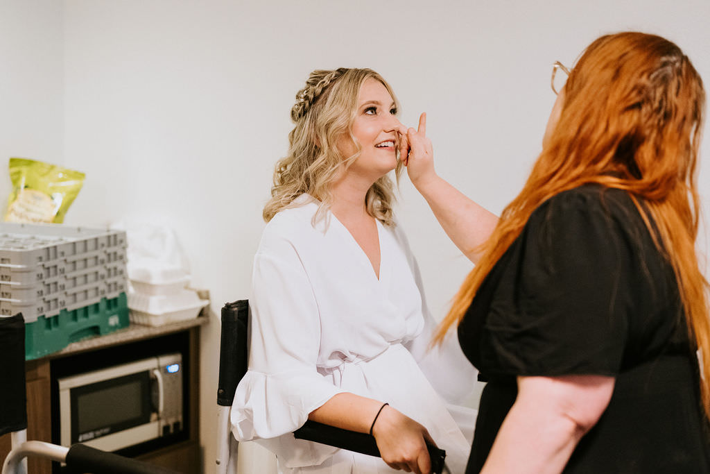 A makeup artist applying makeup to a bride's face as she sits in a chair. 