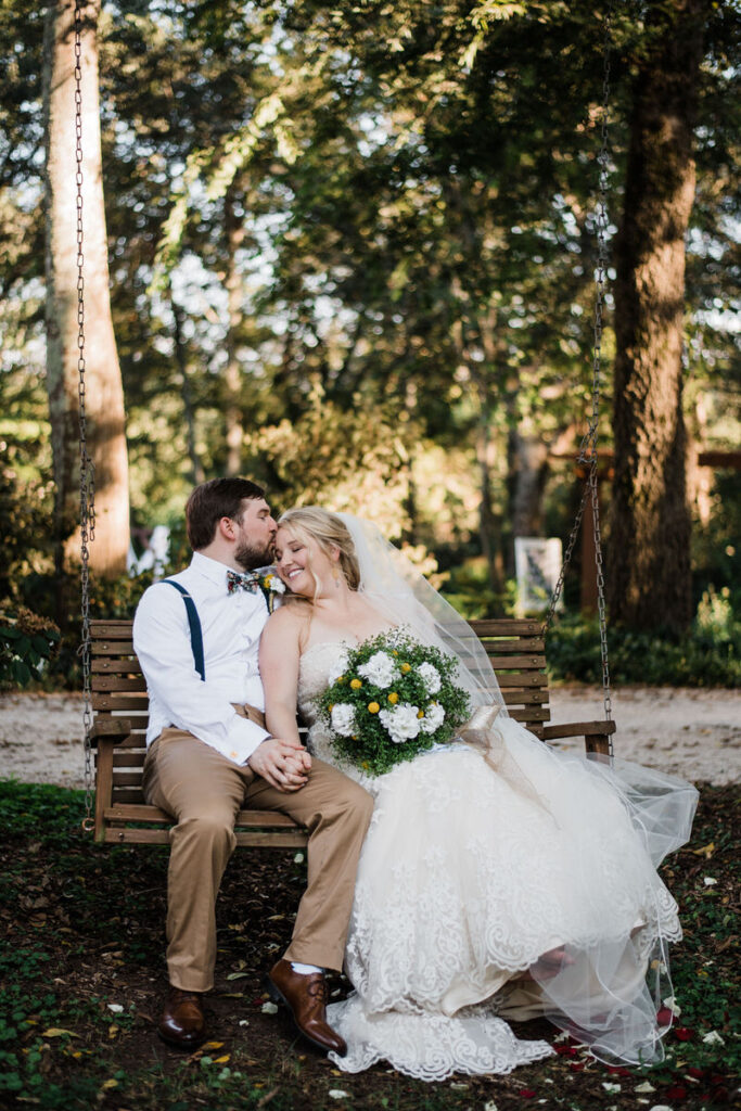A wedding couple holding hands and sitting on a wooden swing while one kisses the other's head.