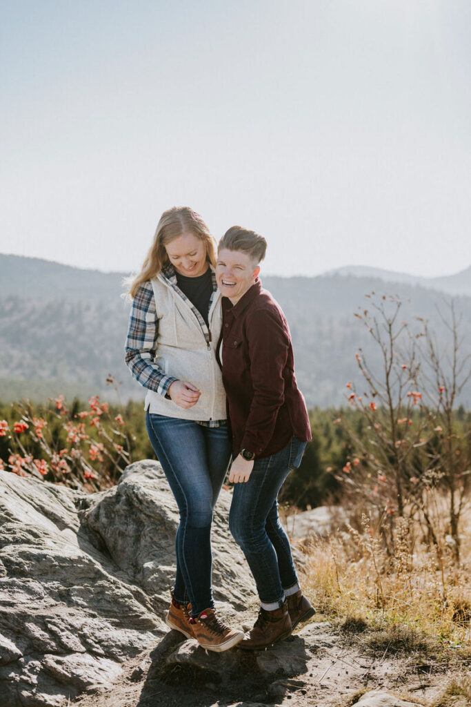 A joyful couple smiling and hugging, standing on a rocky terrain with a blurred natural landscape in the sunshine behind them.