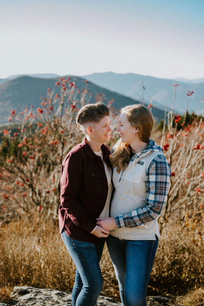 A couple facing each other, sharing a smile, with mountains and red autumn bushes in the soft-focus background.