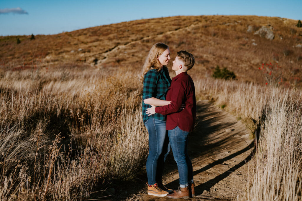 A couple standing close, embracing on a dirt path amidst tall, dry grass with the sun setting behind them, illuminating the scene in golden light.