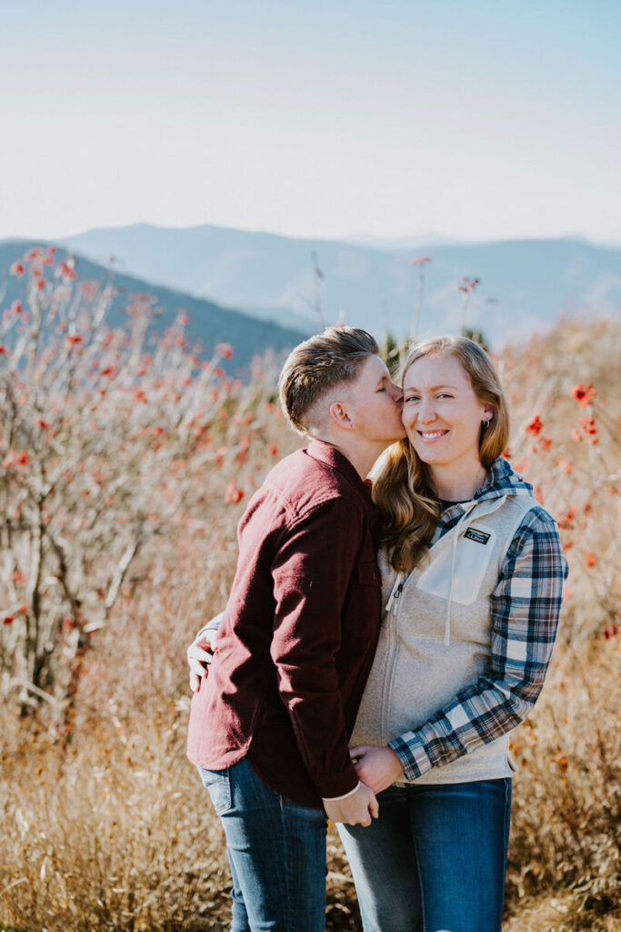 A person kissing their partner on the cheek, both smiling gently, against a backdrop of vibrant autumn shrubs and distant mountains.
