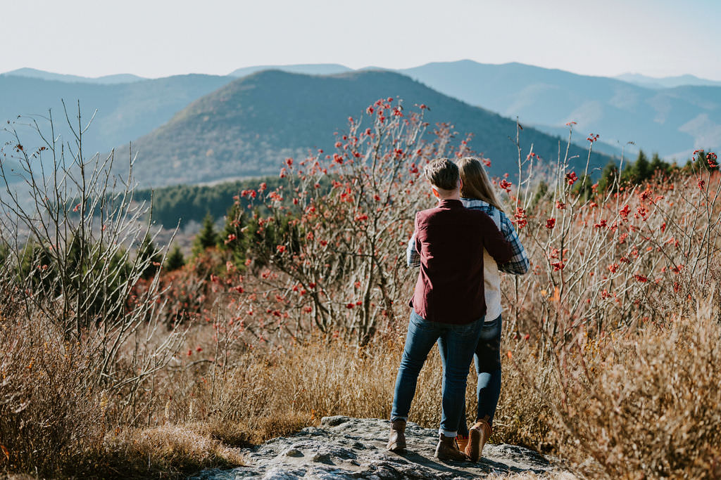 A couple standing from behind, overlooking a mountainous landscape with dry winter shrubs and red berry bushes in the foreground during their Black Balsam Knob engagement photos.