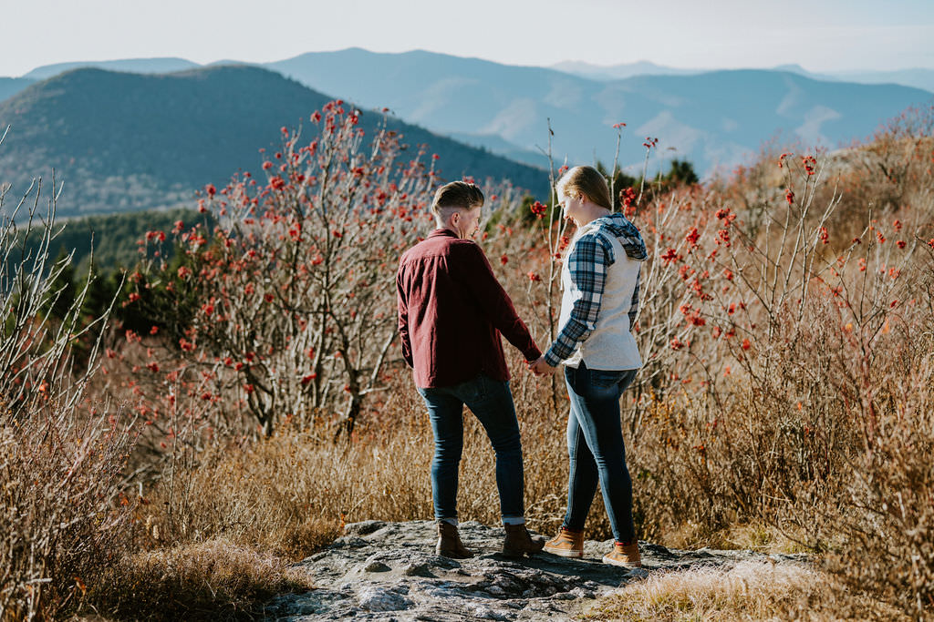 A couple holding hands, facing away, on a rocky outcrop with a backdrop of mountains and autumnal foliage.
