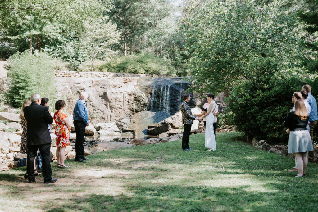 A couple standing up saying their vows in a garden area with a few friends watching them. 
