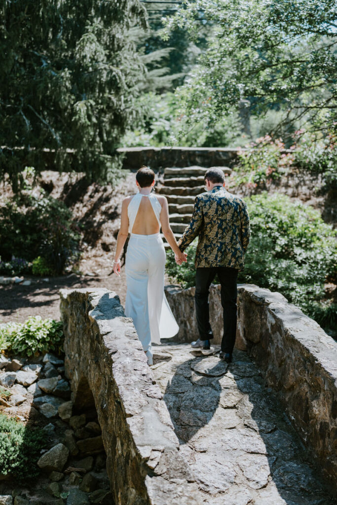 A couple holding hands and walking over a small stone bridge. 
