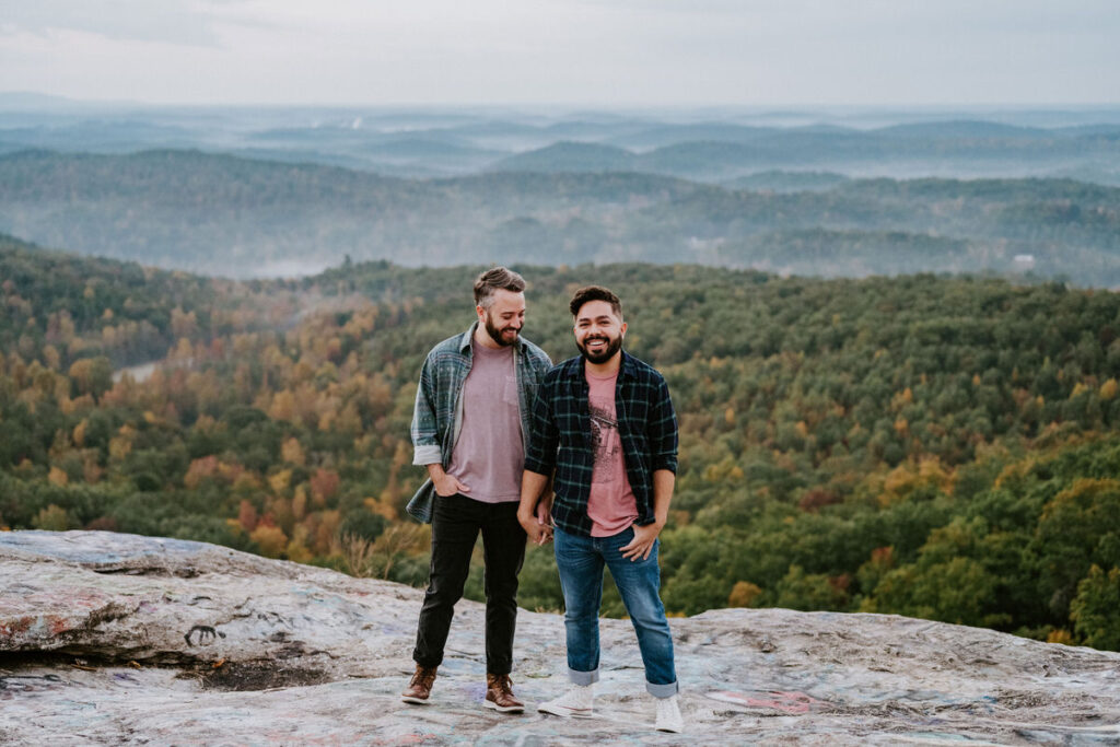 A couple holding hands and laughing while standing on an overlook in the mountains. 
