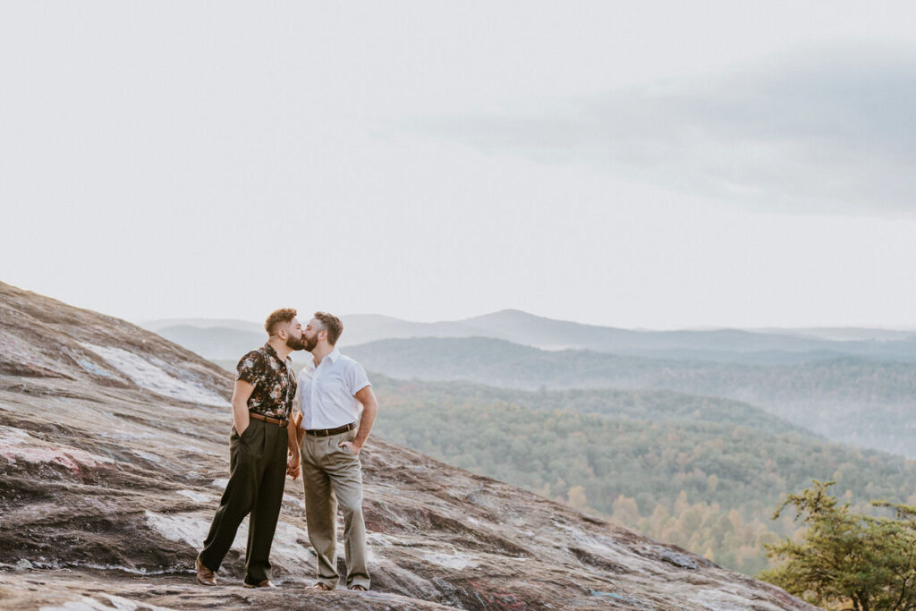 A couple kissing while standing on a rock overlooking the mountains. 
