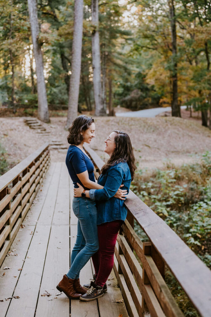 A couple with their arms around each other's waist as they lean up on the railing of a small walking bridge. 
