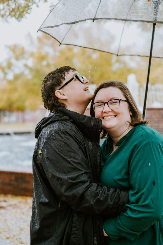 A cheerful couple under a transparent umbrella, the person on the left looking up joyfully as raindrops speckle their jacket, while the person on the right, in glasses and a green blouse, smiles towards the camera