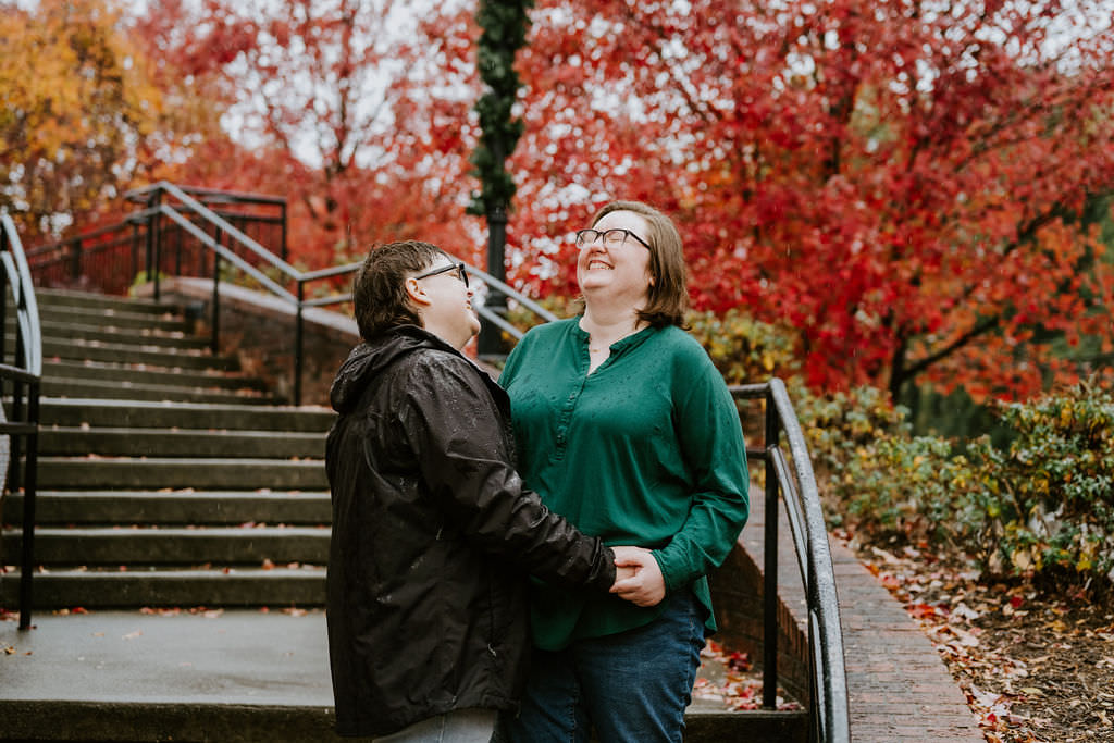 Two individuals stand laughing amidst a vibrant autumnal scene, with one looking at the other affectionately, as they share a warm embrace under a light rain