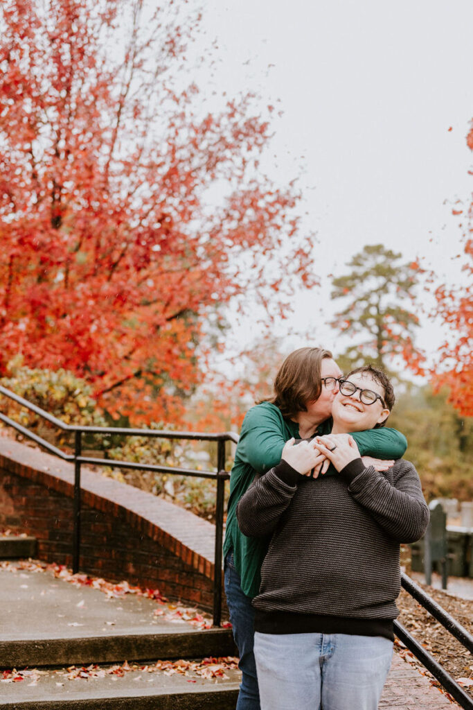 The same couple, framed by the vivid reds and oranges of fall foliage, share a tender moment, one kissing the other's forehead as they hold hands