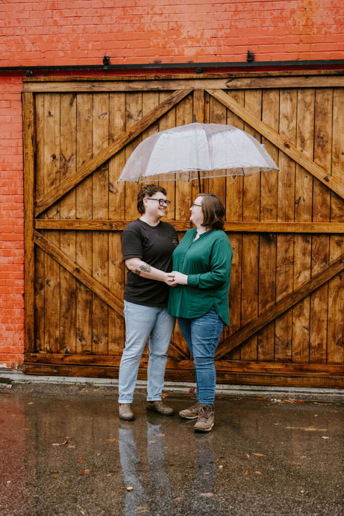 A couple stands close to each other, holding a transparent umbrella on a rainy day, in front of a large wooden door. The warm smile they share contrasts with the cool, wet weather
