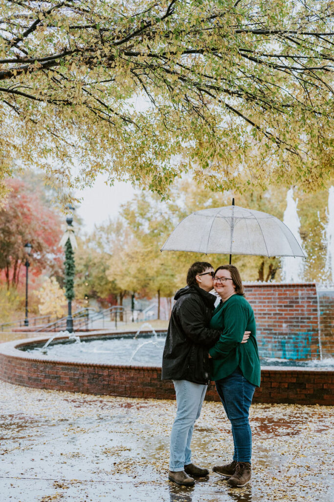 A couple embraces under a clear umbrella amidst a rain shower, with vibrant autumn leaves and a fountain in the background, sharing a kiss and a joyful moment