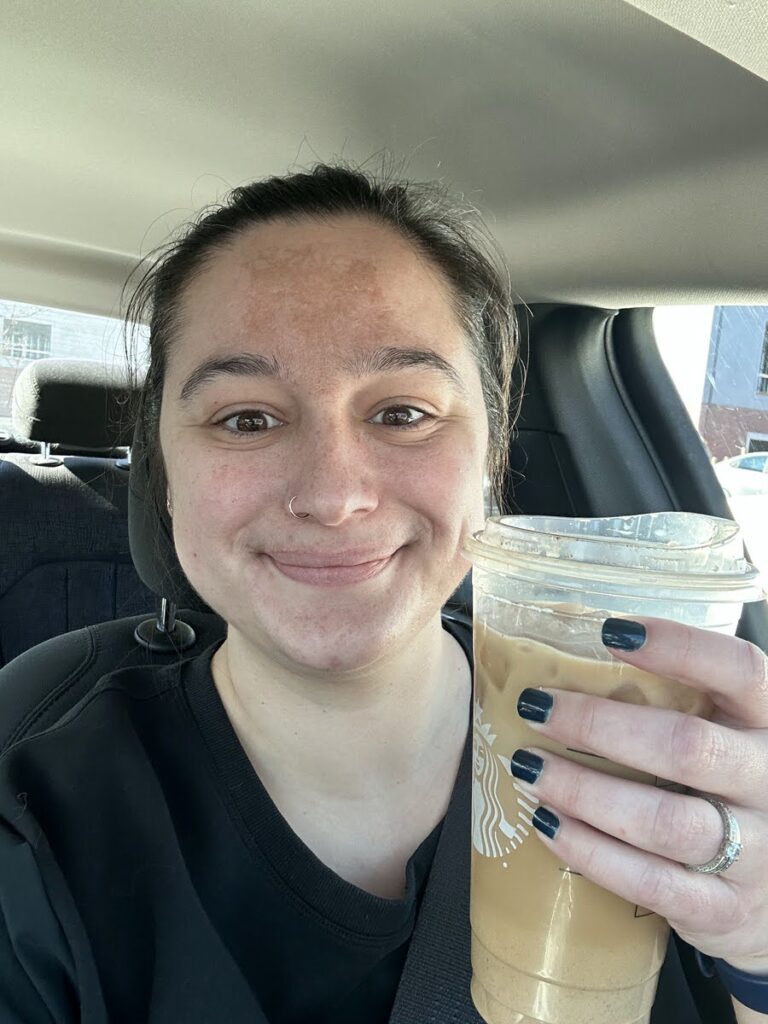 A smiling selfie of a woman enjoying a Starbucks iced coffee in her car, showcasing a casual moment in her day.