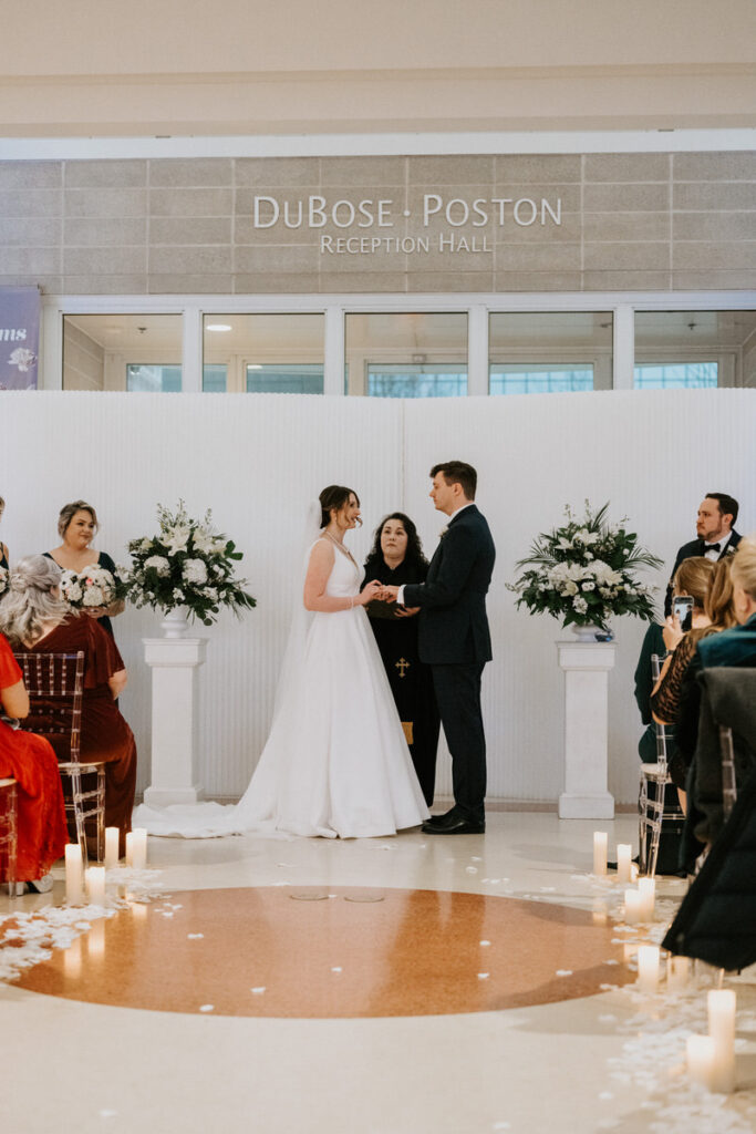 A wedding ceremony in progress at the 'Dubose Poston Reception Hall', with the bride and groom exchanging vows in front of a white backdrop adorned with floral arrangements and surrounded by guests.