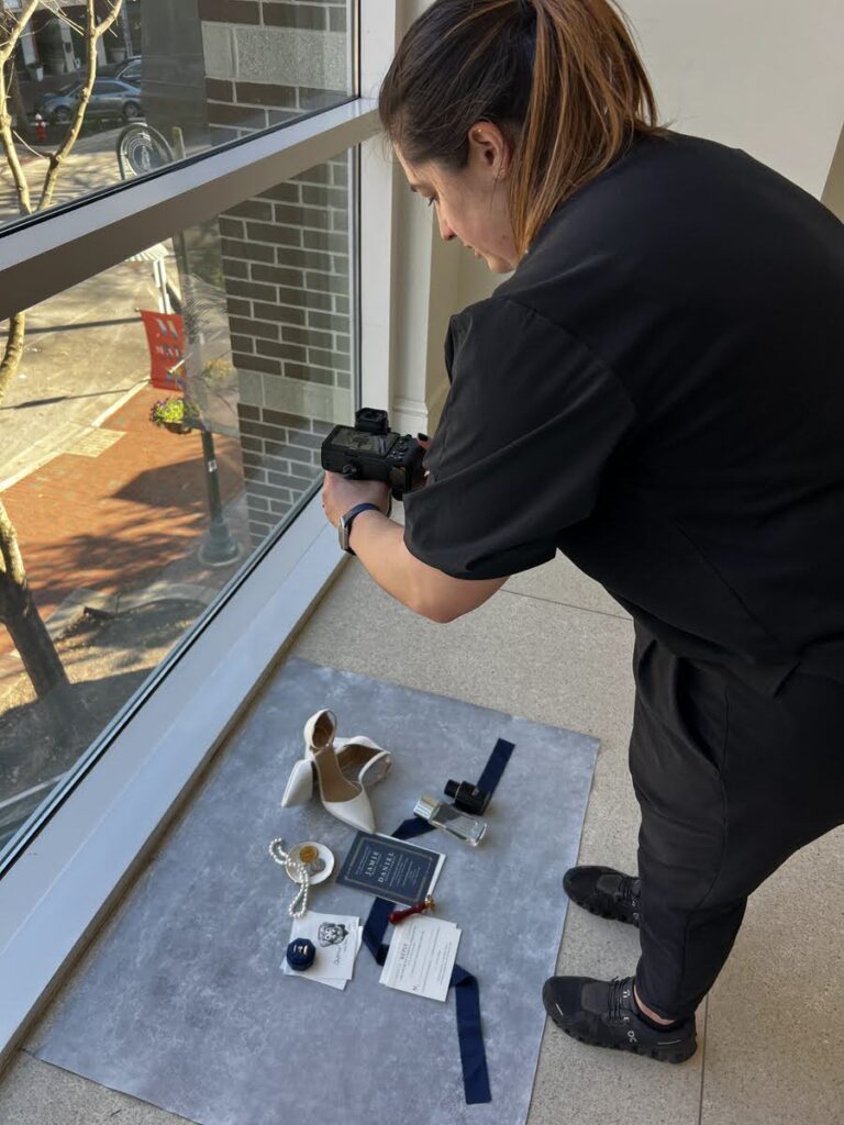 A photographer crouched by a window, capturing detailed shots of wedding items spread out on the floor, showcasing the meticulous process of wedding photography.