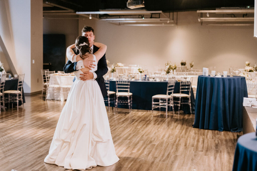 The newlyweds dancing alone in a large reception hall, sharing a tender embrace, with tables and chairs in the background signifying the end of the wedding celebration.