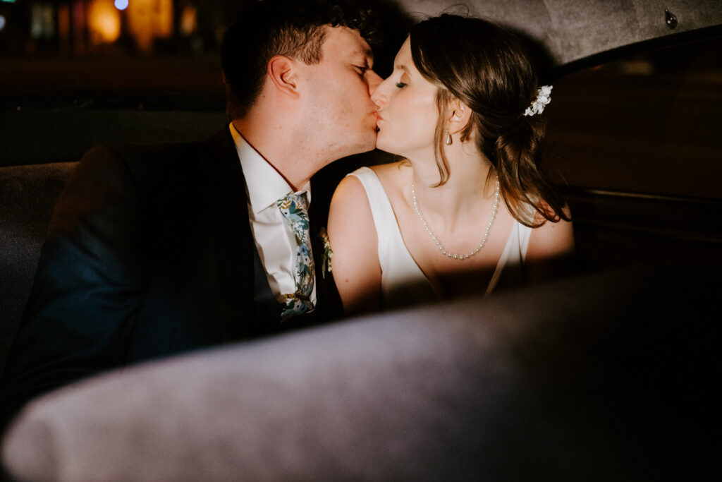 A candid shot inside a dimly lit vehicle showing the bride and groom sharing an intimate kiss, capturing the privacy and romance at the end of their wedding night.