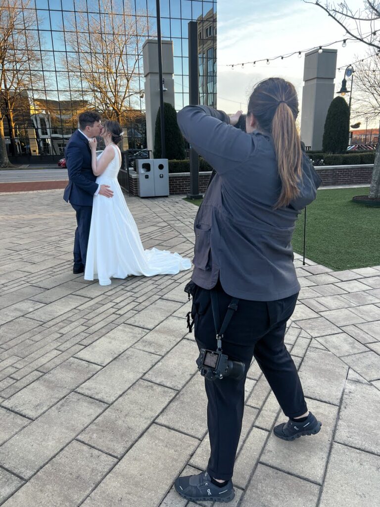 A photographer taking formal wedding photographs of a bride and groom embracing each other, set against an urban backdrop with a reflection of the sunset.
