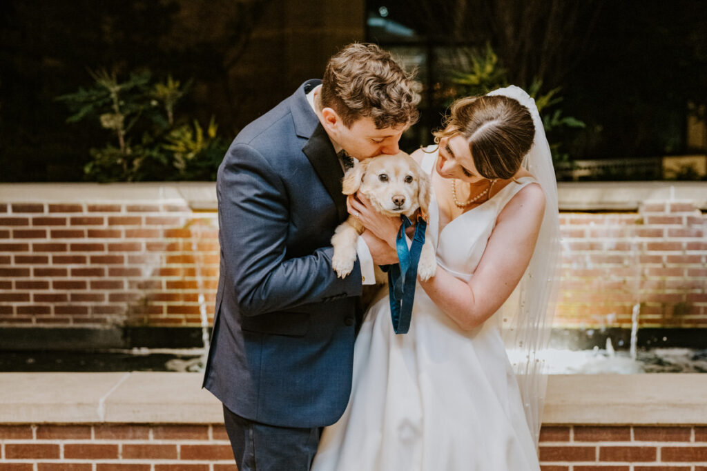 A close-up wedding photo capturing a tender moment as the bride and groom lovingly kiss their puppy, set against the backdrop of a brick water fountain.