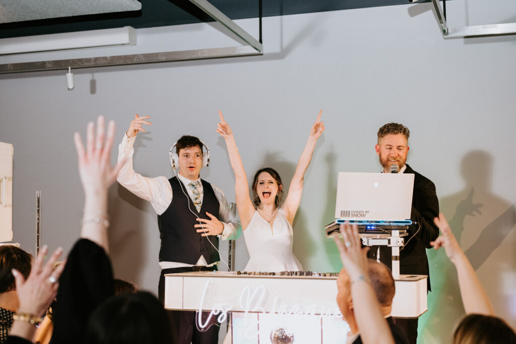 A lively scene from a wedding reception with the bride and groom at the DJ booth, celebrating with guests, hands in the air, during the party.