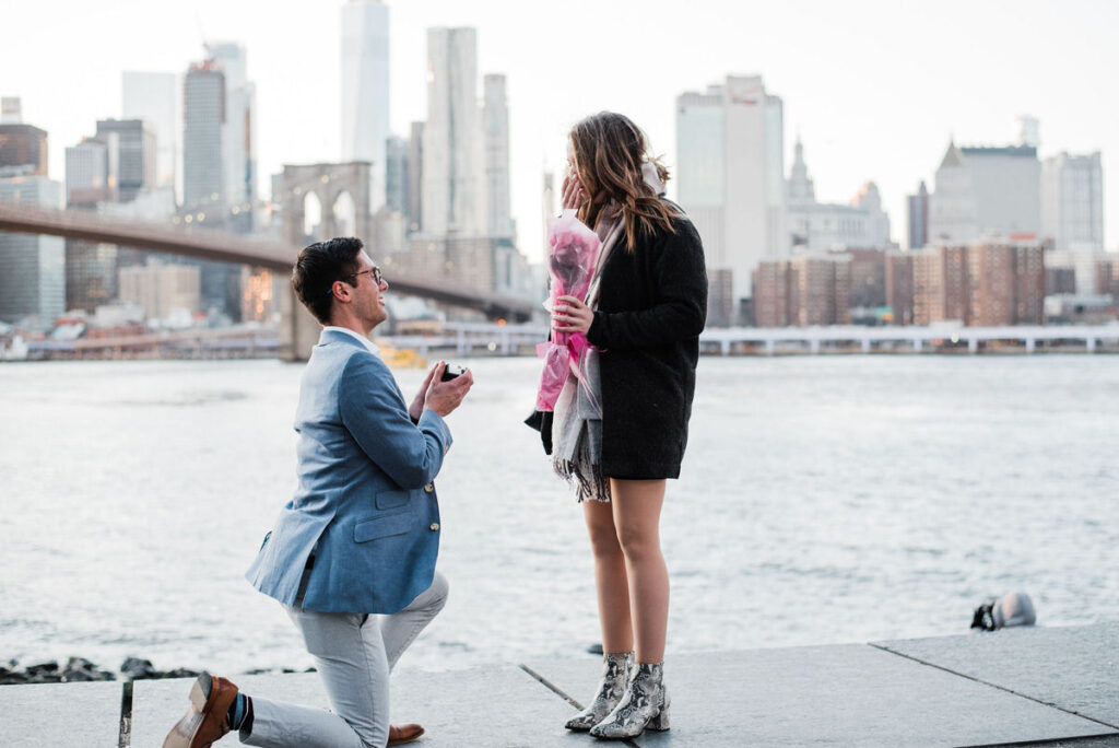 Man in a blue blazer and grey pants kneeling with a ring box, proposing to a woman in a black coat holding pink roses, with the cityscape in the background