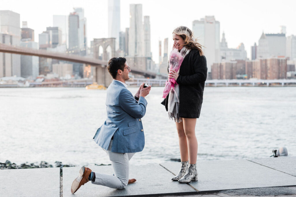 Couple engaged in a joyous proposal by the waterfront, with the Brooklyn Bridge in the background during twilight