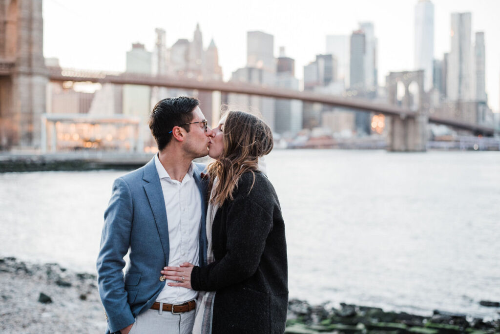 Man and woman kissing on the waterfront with the Brooklyn Bridge and New York City skyline in the soft light of dusk