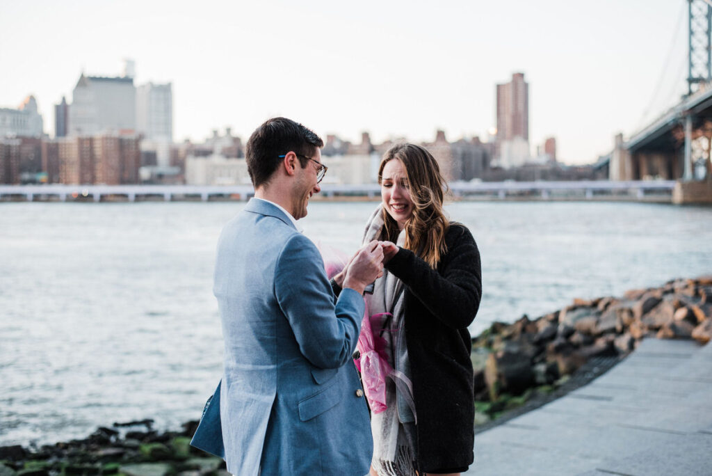 Couple standing close, holding hands and smiling, with the Brooklyn Bridge and city skyline illuminated by the soft glow of sunset