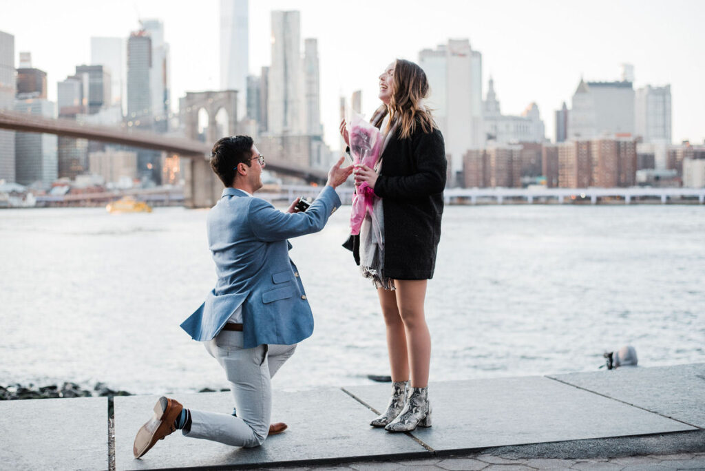 Woman in a black coat and boots, smiling down at a man proposing to her against a backdrop of the Brooklyn Bridge and city skyline