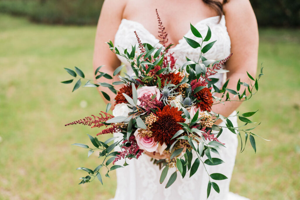 Close-up of a bride's hands holding a vibrant wedding bouquet featuring dark reds, pinks, and lush greenery, with her white lace wedding gown partially visible